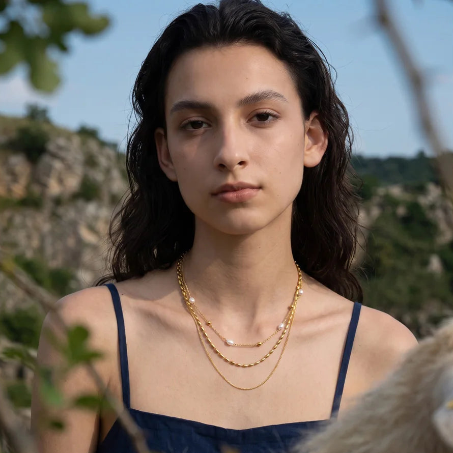 A woman outdoors wearing layered gold necklaces, including a delicate chain with small white pearl-like beads, over a navy blue top. The background shows a blurred natural landscape with greenery and rocks.