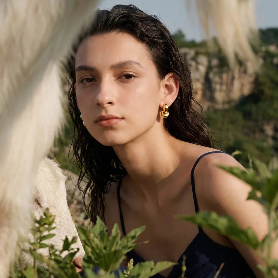 A woman outdoors wearing a small gold hoop earring, with a blurred animal and greenery in the foreground and a rocky landscape behind her.